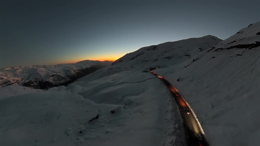 An aerial view shows a line of cars with their lights on driving along a winding road through a snowy mountain pass at dusk