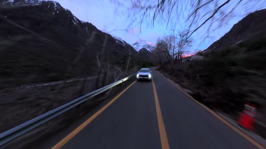 A white station wagon drives along a winding mountain road at dusk, with a vibrant sunset illuminating the snow-capped peaks in the distance