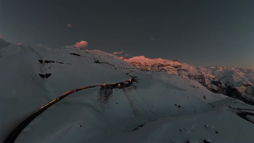 Aerial time-lapse of cars with headlights driving on a winding road through a majestic, snow-covered mountain range at sunset