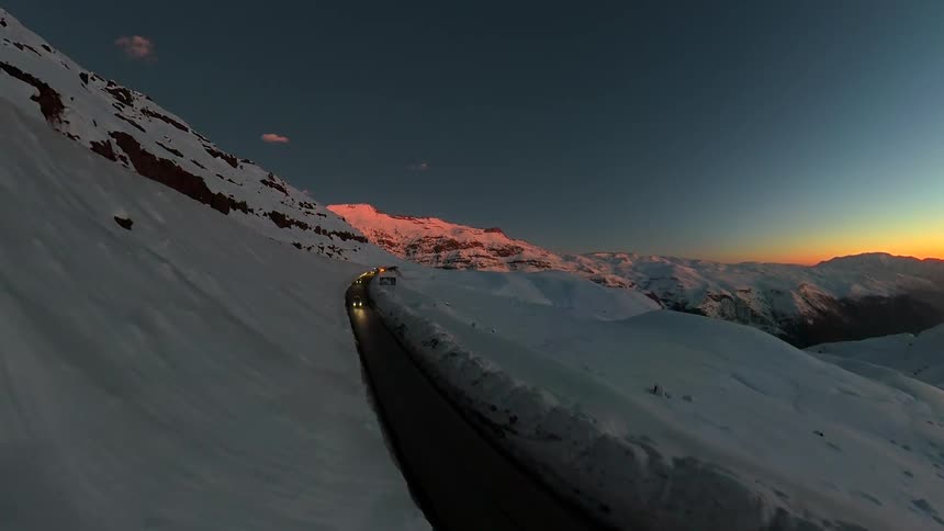 An aerial drone follows a winding road through a snow-covered mountain pass as cars drive with their headlights on during a beautiful sunset