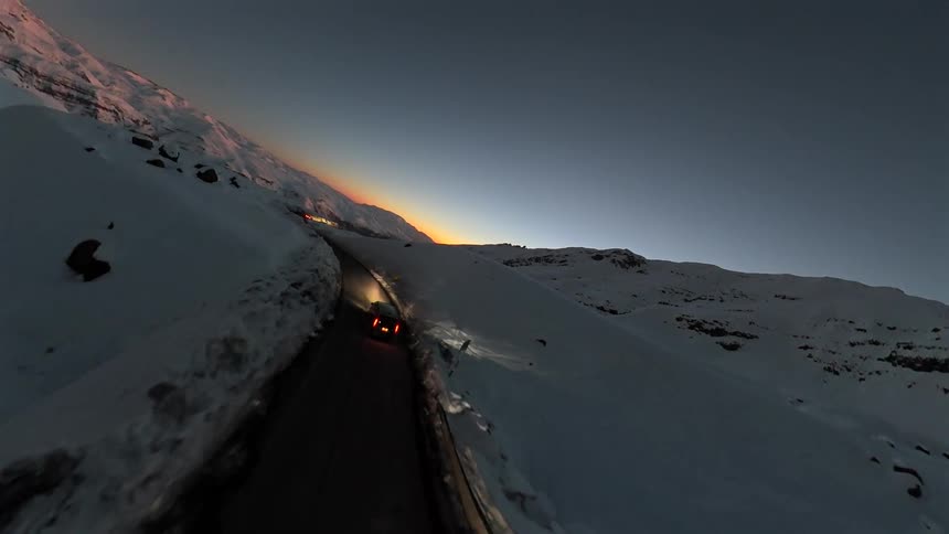 An immersive aerial view follows cars driving along a narrow mountain pass covered in deep snow during a spectacular sunrise