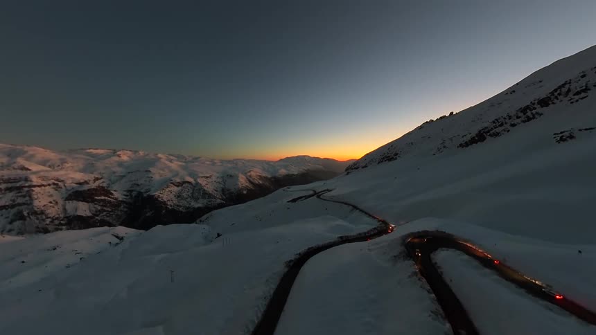 An aerial time-lapse captures the light trails of cars winding through a snowy mountain pass at dusk