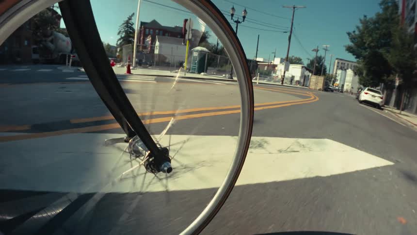A close-up point-of-view shot of a bicycle's front wheel spinning as it rides through city streets on a sunny day