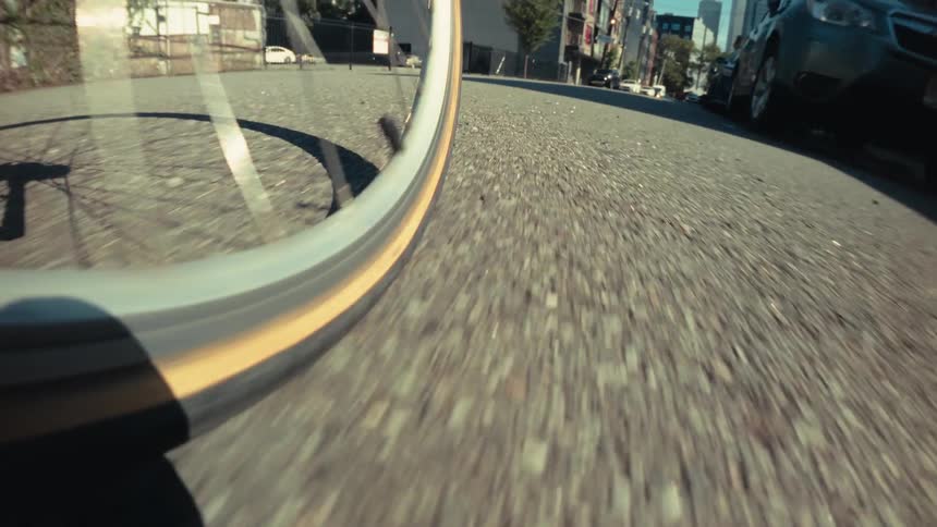 A slow-motion, low-angle point-of-view shot captures a bicycle wheel rolling down a sunny city street and coming to a stop