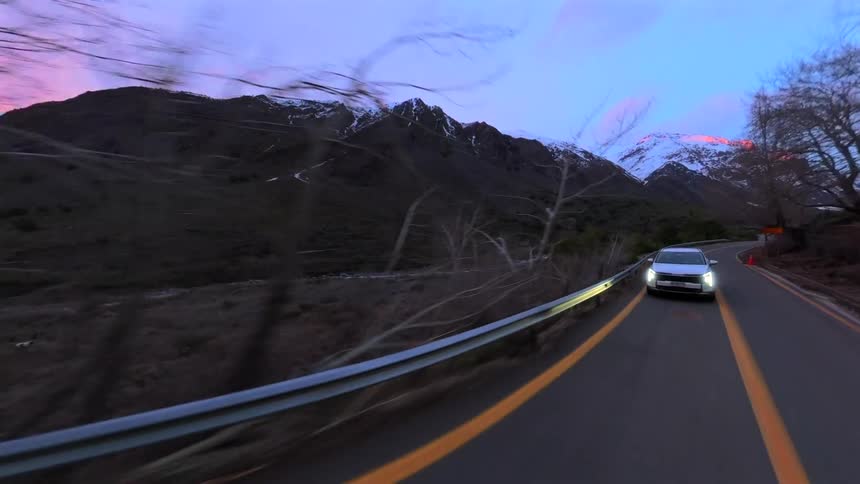 A car drives on a winding road through a majestic mountain valley with snow-capped peaks during a vibrant sunrise