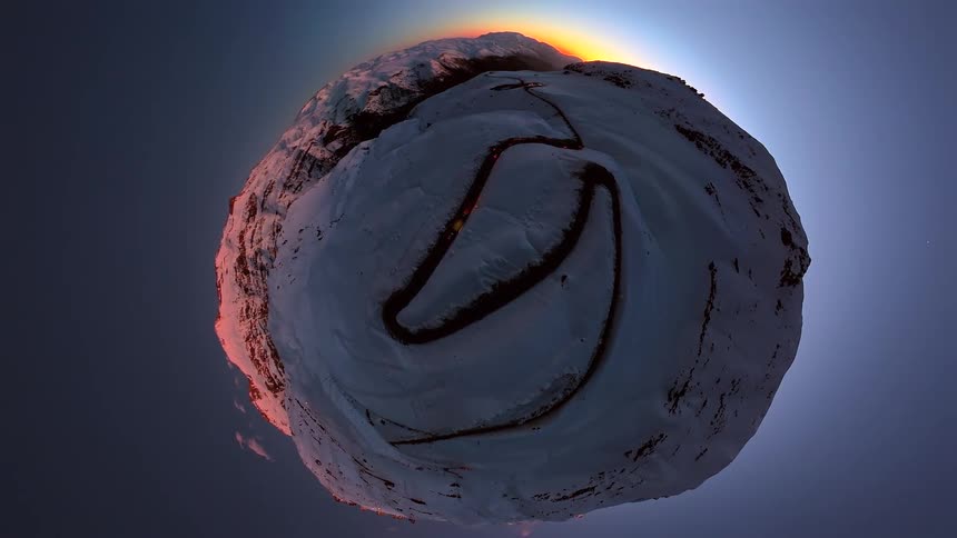 A surreal tiny planet view of a lone car with headlights on driving up a winding road on a snowy mountain at sunset