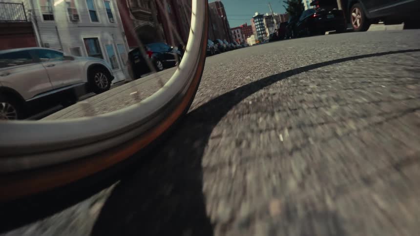 A low-angle point-of-view shot follows a bicycle wheel as it rolls down a sunlit city street lined with parked cars
