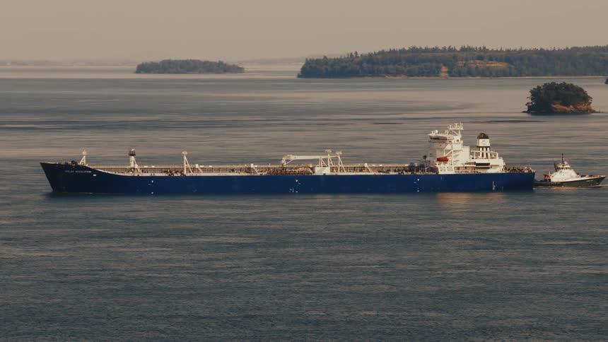 A large oil tanker is escorted by a tugboat as it sails through calm waters past tree-covered islands