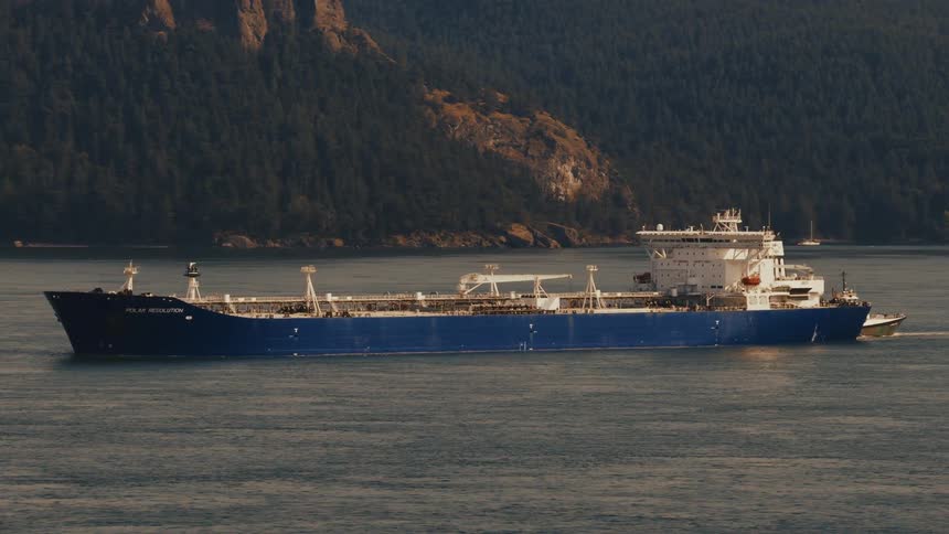 A large blue oil tanker named Polar Resolution sails across the water, guided by a tugboat, with a forested coastline in the background