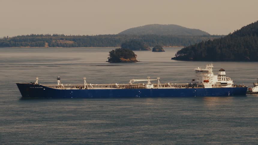 A tugboat guides a large oil tanker through a calm channel surrounded by lush, green islands