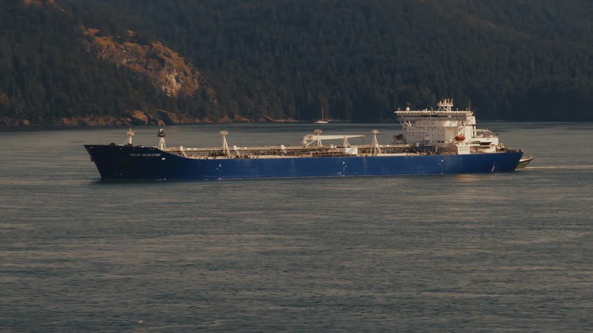 A large blue oil tanker named Polar Resolution sails on the calm ocean waters past a tree-covered shoreline