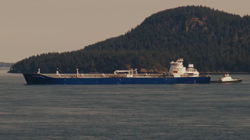 A large blue oil tanker is escorted by a tugboat past a lush green, tree-covered island