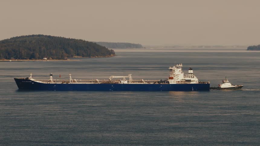 A large blue oil tanker is pushed by a tugboat through a channel with forested islands in the background