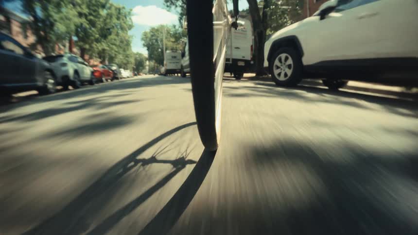 Point-of-view shot of a bicycle's front wheel and its shadow spinning down a sunny city street lined with parked cars and trees