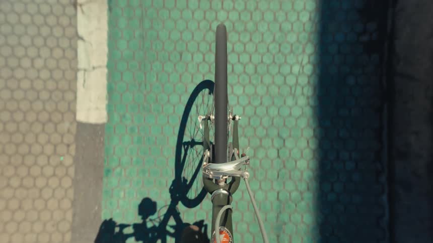 A point-of-view shot from above shows the front wheel of a bicycle rolling along a green bike lane and then onto the asphalt street on a sunny day
