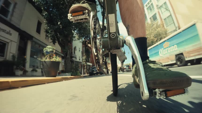 A dynamic low-angle point-of-view shot of a person's feet pedaling a bicycle down a sunny city street