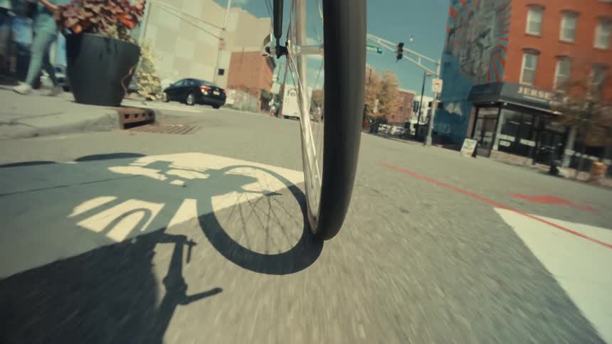 A low-angle point-of-view shot of a bicycle's front wheel rolling down a sunny city street, passing parked cars and casting a shadow on the asphalt
