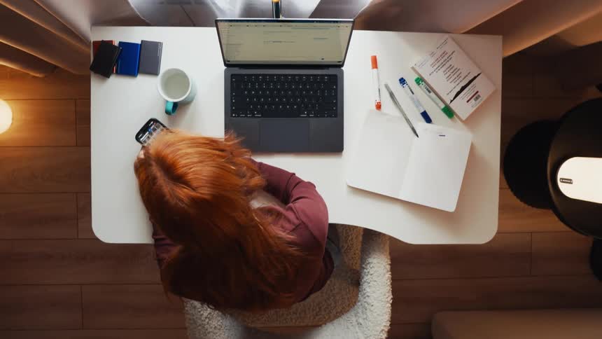 Top-down time-lapse of a busy redhead working at her desk, switching between her laptop, notebook, and phone