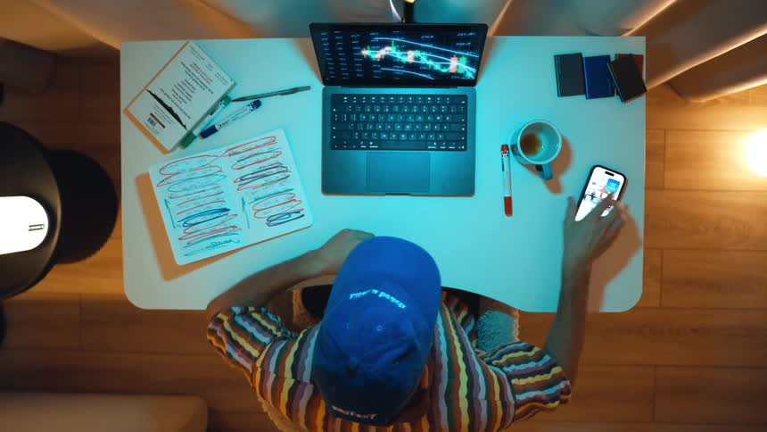 Top-down vertical shot of a man in a blue cap analyzing stock market charts on his laptop and smartphone while working from home at night