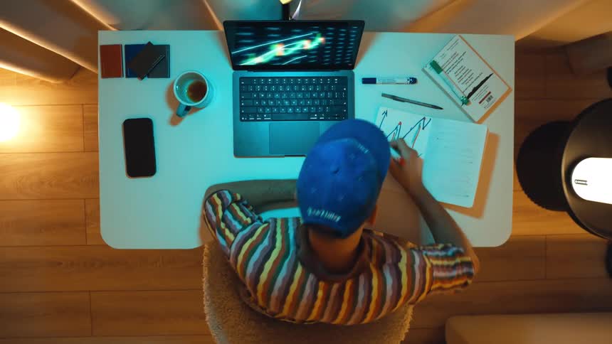 Top-down timelapse of a male trader working late at home, analyzing stock market charts on his laptop and in a notebook