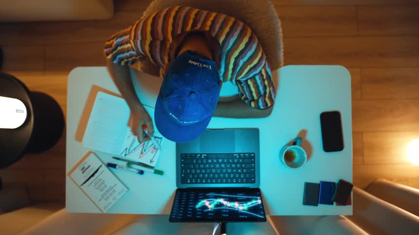 Overhead view of a man in a cap analyzing stock market data on a laptop and drawing charts in a notebook at night