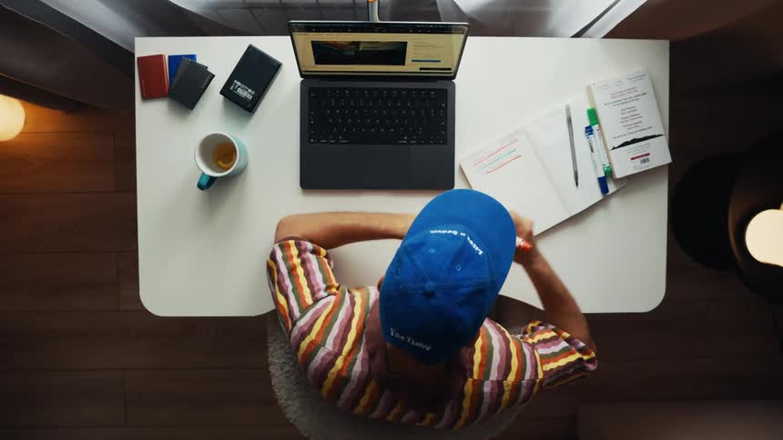 A top-down view of a man in a blue cap working from home, taking notes in a notebook while using a laptop and drinking from a mug