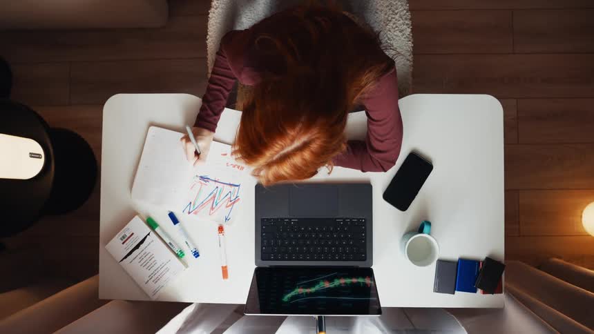 Top-down timelapse of a frustrated female trader analyzing falling stock market charts on her laptop and taking notes at night