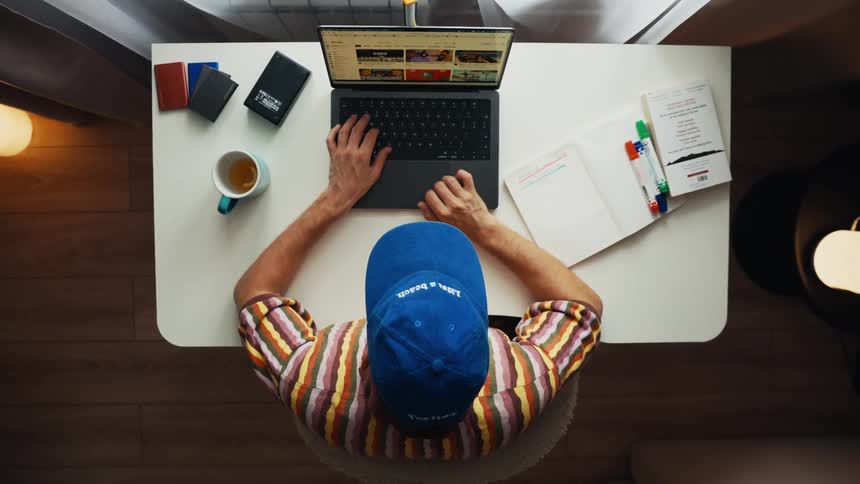 Top-down view of a man working from home, taking notes in a planner, and using a laptop at his desk