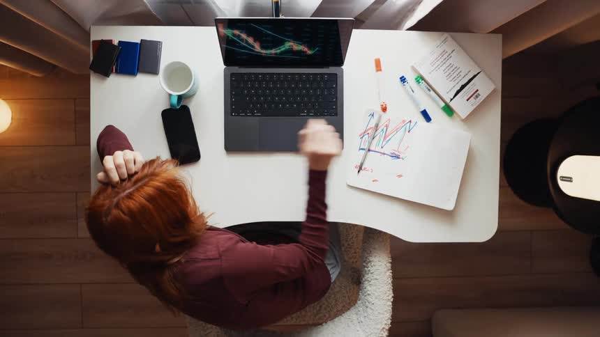 Top-down view of a woman with red hair trading stocks from her home office, analyzing charts on her laptop and drawing in a notebook