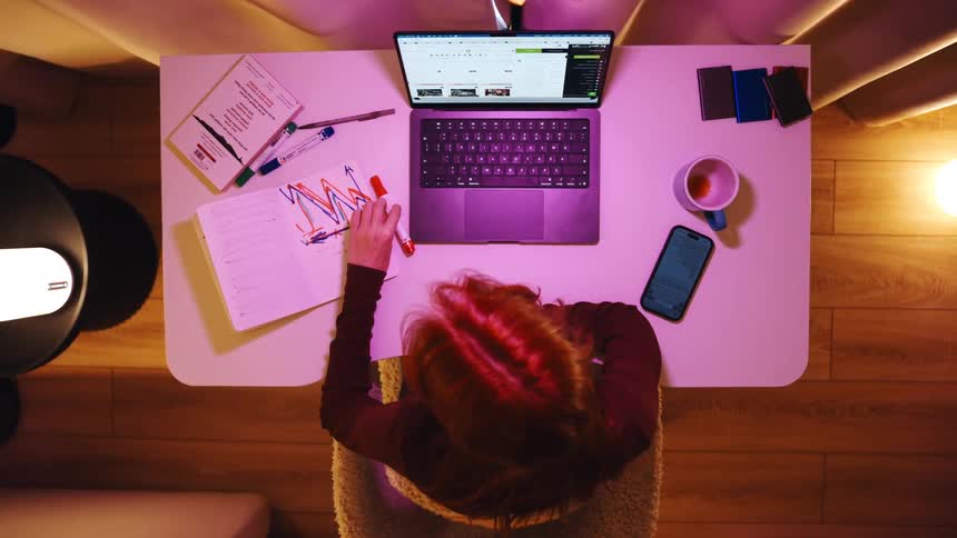 Top-down view of a woman working from home at night, multitasking between her laptop, phone, and notebook with charts