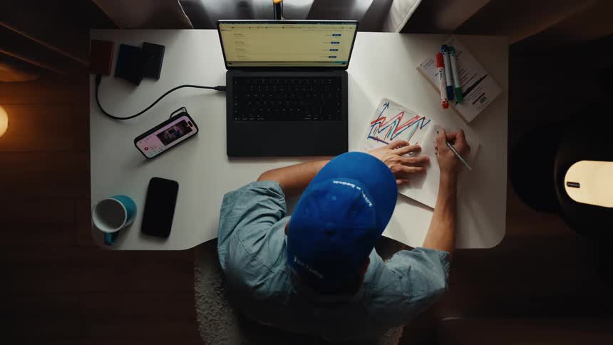 Top-down view of a man in a blue cap working from his home office, taking notes and drawing charts in a notebook next to his laptop