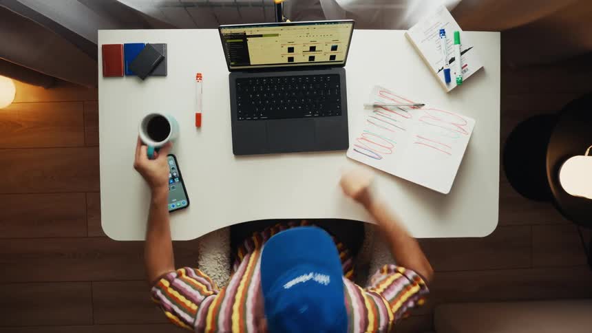 Overhead time-lapse captures a busy freelancer in a blue cap multitasking between a laptop, notebook, and phone at a modern home office desk