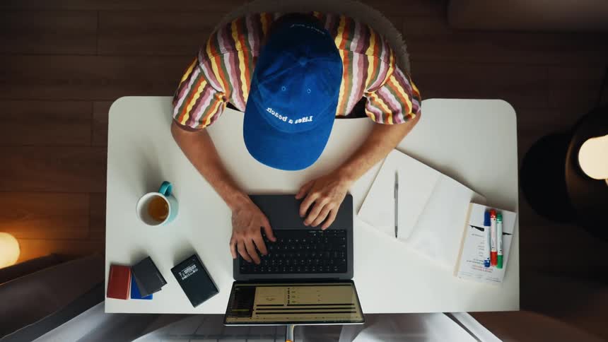 A top-down view of a man in a blue cap multitasking as he types on his laptop and takes notes in a notebook at his desk