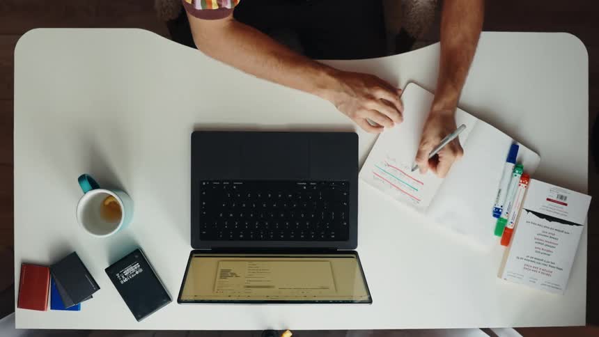 Top-down view of a person typing on a laptop and taking notes in a notebook at a modern white desk