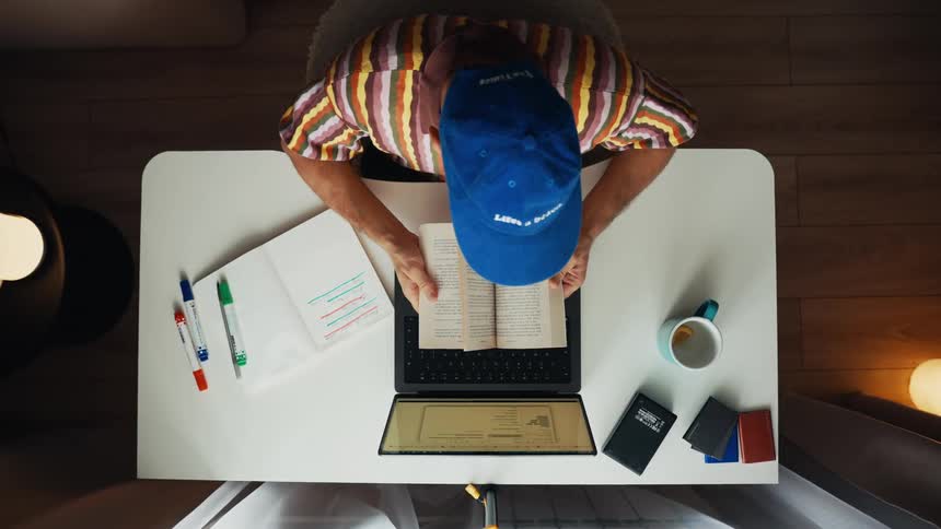 Top-down view of a person in a colorful shirt and blue cap studying at a white desk, opening a book, and reading