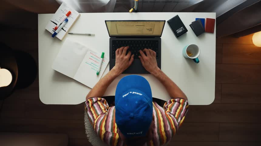 A top-down time-lapse of a man in a blue cap multitasking at his desk, working on a laptop, reading a book, and writing in a notebook