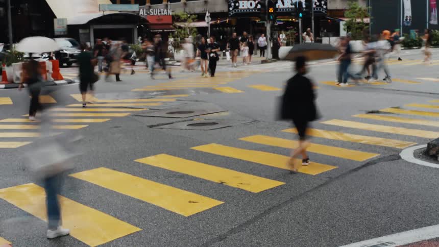 A dynamic long-exposure time-lapse shows the blurred motion of cars and crowds of people at a bustling city intersection