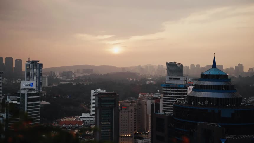 A modern city skyline surrounded by lush green hills is bathed in the warm, hazy light of a setting sun