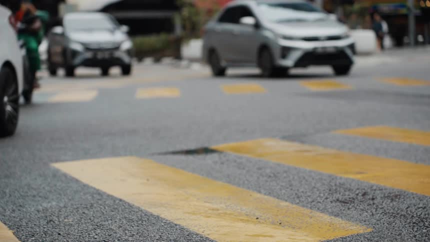A slow-motion, out-of-focus shot of cars driving and people crossing a city street with yellow road markings in the foreground