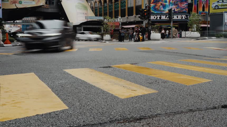 A low-angle time-lapse shows motion-blurred traffic speeding through a busy city intersection with a yellow crosswalk in the foreground