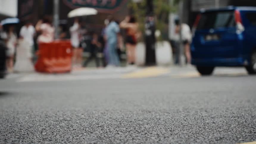 A low-angle view shows the lower half of cars driving down a busy city street, with the asphalt in focus and pedestrians blurred in the background