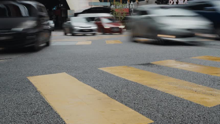 A dynamic low-angle time-lapse captures blurred traffic speeding through a busy city intersection with a yellow crosswalk in the foreground