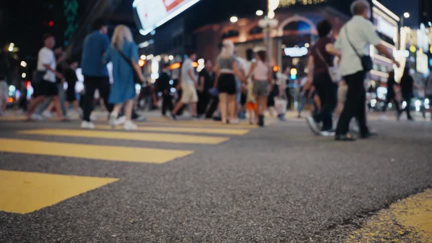 A low-angle, out-of-focus shot of a crowd of pedestrians' feet as they walk across a yellow-striped crosswalk in a bustling city at night