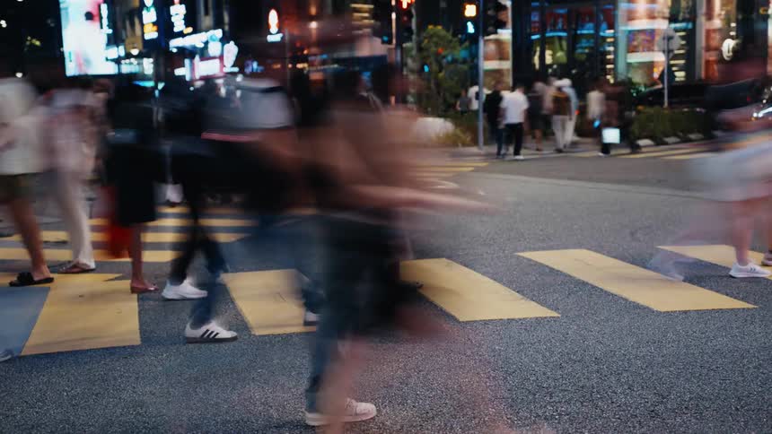 Timelapse footage captures the motion blur of a crowd walking across a yellow crosswalk in a bustling city at night