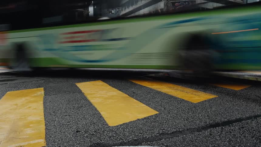 A low-angle time-lapse captures the blur of traffic as cars, buses, and trucks speed over a yellow crosswalk at a busy city intersection