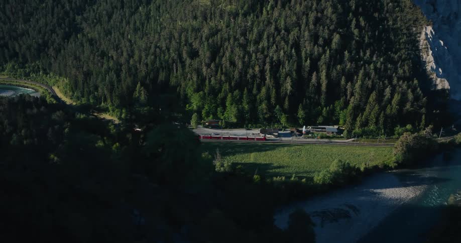 An aerial view shows a red passenger train traveling through a lush green mountain valley alongside a river in the Swiss Alps