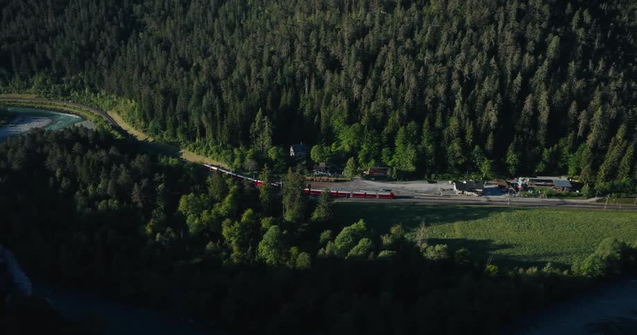 A high aerial shot captures a red passenger train moving through a beautiful, sunlit green valley next to a dense forest and a river