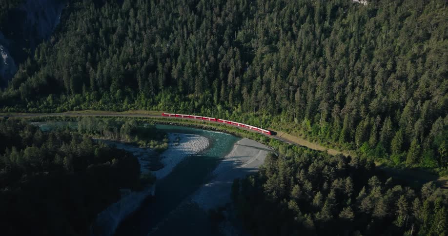 An aerial view of a red passenger train traveling through a dense green forest alongside a scenic river