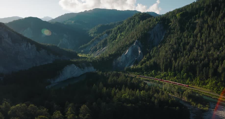 A scenic red train travels through a stunning, sunlit mountain valley covered in lush green forests alongside a river