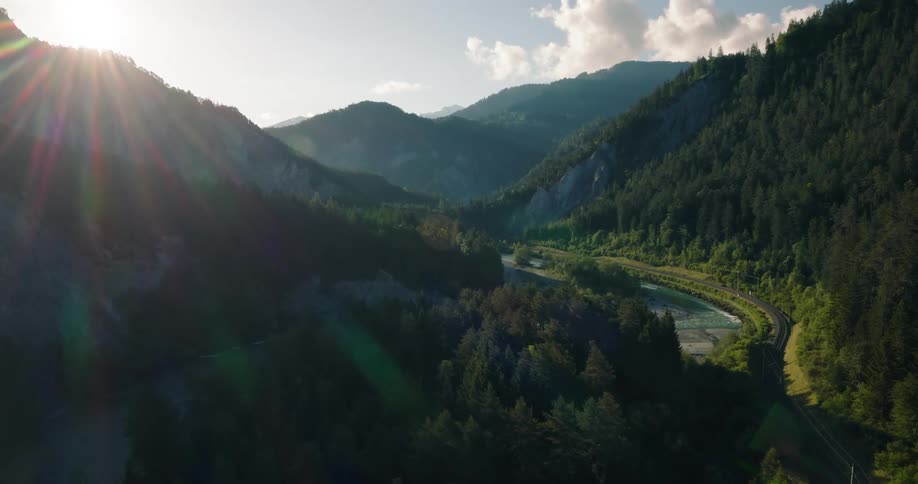 Expansive aerial pullback shot of the sun shining over a lush green mountain valley with a river and train tracks